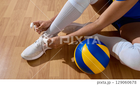Athlete lacing shoes beside volleyball on wooden court surface 129060678