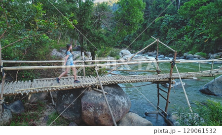 Aerial drone view capturing a female tourist enjoying a hike while crossing a small wooden bridge over a flowing river, surrounded by lush tropical forests in Southeast Asia Aerial drone view capturing a female tourist enjoying a hike while crossing a small wooden bridge over a flowing river, surrounded by lush tropical forests in Southeast Asia 129061532