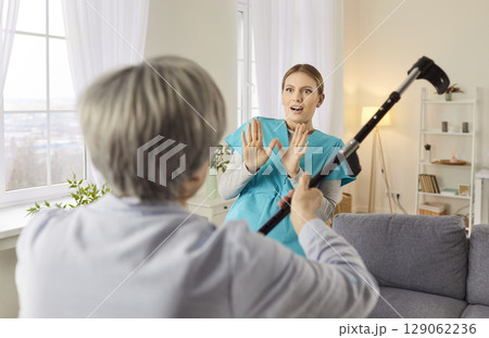Angry elderly senior woman going to hit young female nurse with crutch. Angry elderly senior woman going to hit young female nurse with crutch. 129062236