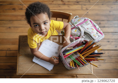 Young child with curly hair studying at a wooden table surrounded by colorful school supplies Young child with curly hair studying at a wooden table surrounded by colorful school supplies 129062720
