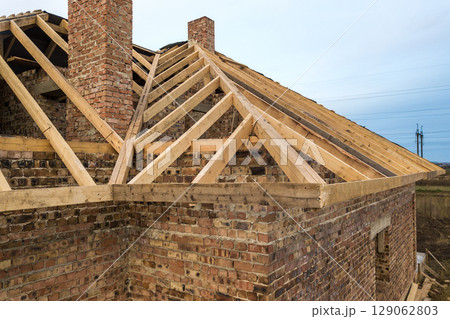 Private residential house with wooden roof frame structure under construction. Unfinished brick building under development. 129062803
