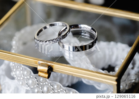 Wedding rings and garter laying in a glass box with white roses in the background 129065116