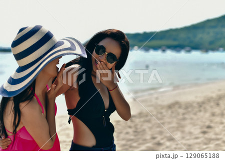Two friends enjoying a sunny day at the beach, sharing laughter and smiles while dressed in vibrant beachwear. 129065188