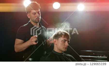 Young Man with Beard at Barber Shop. Barber is Preparing his Haircut. Dark Atmosphere, Black Background. Concept of Barber and a Client. Young Man with Beard at Barber Shop. Barber is Preparing his Haircut. Dark Atmosphere, Black Background. Concept of Barber and a Client. 129065199