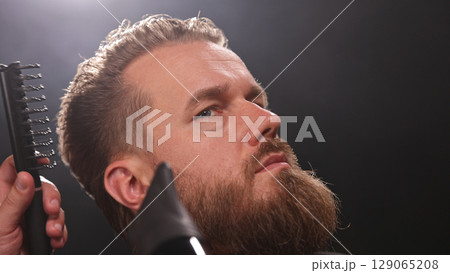 Young Man with Beard at Barber Shop. Barber is Preparing his Haircut. Dark Atmosphere, Black Background. Concept of Barber and a Client. 129065208
