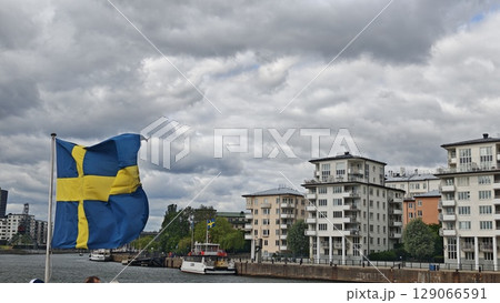 Swedish flag waving in the wind by the waterfront with boats and modern buildings in the background Swedish flag waving in the wind by the waterfront with boats and modern buildings in the background 129066591