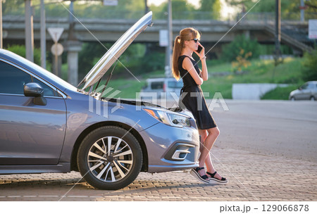 Helpless woman standing near her car with open hood takling on mobile phone calling road service for help. Young female driver having trouble with vehicle 129066878