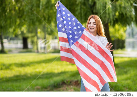 Happy young woman posing with USA national flag standing outdoors in summer park. Positive girl with United States banner outdoors. Happy young woman posing with USA national flag standing outdoors in summer park. Positive girl with United States banner outdoors. 129066885