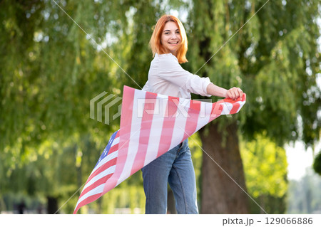 Happy young red haired woman posing with USA national flag standing outdoors in summer park. Positive girl celebrating United States independence day. Happy young red haired woman posing with USA national flag standing outdoors in summer park. Positive girl celebrating United States independence day. 129066886