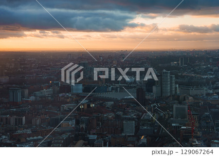 The London Eye stands prominently against a sunset sky, with clouds adding depth. The cityscape features diverse architecture, capturing London's urban landscape. 129067264