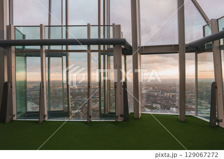 The Shard's modern interior features large glass panels and green turf flooring, offering a panoramic view of London's cityscape under a cloudy sky. 129067272