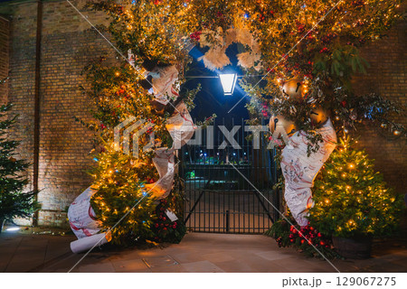A decorated archway with golden lights, greenery, and baubles against a brick wall. A wrought iron gate leads to a softly lit area beyond. 129067275