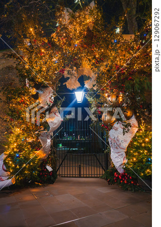 A decorated archway with golden bells, red berries, and greenery, illuminated by a street lamp. A wrought iron gate suggests a park entrance in London. A decorated archway with golden bells, red berries, and greenery, illuminated by a street lamp. A wrought iron gate suggests a park entrance in London. 129067292