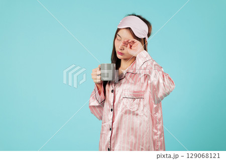 Asian woman in pajama holds a cup after a light sleep, wearing a sleep mask, and relaxing at home, enjoying hot milk for a good mood and restful sleep Asian woman in pajama holds a cup after a light sleep, wearing a sleep mask, and relaxing at home, enjoying hot milk for a good mood and restful sleep 129068121