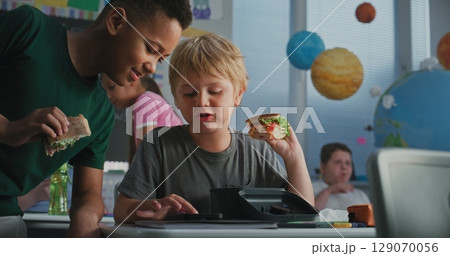 Primary School Boy Sitting at Desk, Eating Sandwich, Using Smartphone and Talking to Classmate 129070056