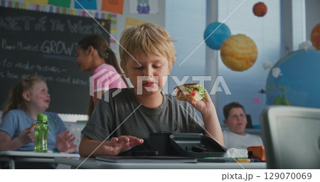 Primary School Boy Sitting at Desk, Eating Sandwich, Using Smartphone and Talking to Classmate 129070069