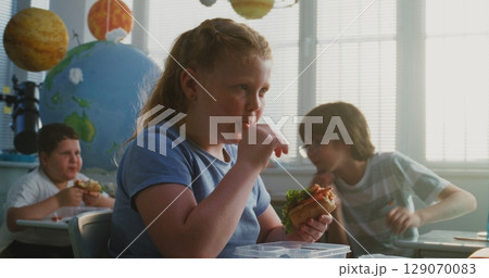 Primary School Girl Eating Delicious Homemade Sandwich from Lunch Box, Talking to Classmates 129070083