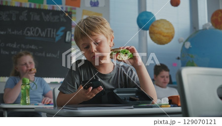 Elementary School Student Eating Sandwich from Lunch Box, Using Phone and Talking to Classmate 129070121