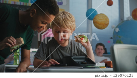 Primary School Boy Sitting at Desk, Eating Sandwich, Using Smartphone and Talking to Classmate 129070123
