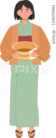 A calm Japanese woman in yukata stands holding a bath bucket and towel. 129076991