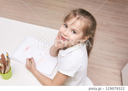 Caucasian female child smiling and drawing at table with colored pencils Caucasian female child smiling and drawing at table with colored pencils 129079322