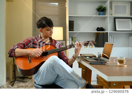 Man playing acoustic guitar while sitting in a modern home office with laptop on the desk 129080876