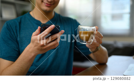 Close up of smiling man holding a smartphone and a glass cup of coffee sitting in living room 129080882
