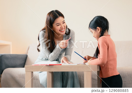 Smiling mother helping her daughter with homework while sitting together at table at home. Smiling mother helping her daughter with homework while sitting together at table at home. 129080885