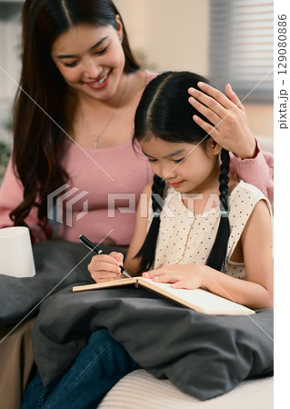 Happy mom supporting her child during homework or creative writing activity on a sofa. Happy mom supporting her child during homework or creative writing activity on a sofa. 129080886