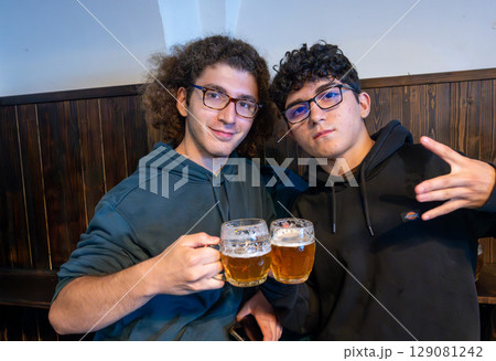 Prague, Czech Republic, August 5, 2023. Two friends look into the camera while toasting in a pub with two mugs of beer. A lifestyle of shared drinking. Prague, Czech Republic, August 5, 2023. Two friends look into the camera while toasting in a pub with two mugs of beer. A lifestyle of shared drinking. 129081242