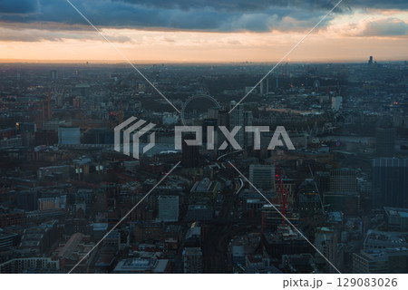 A panoramic view of London featuring the London Eye at sunset. The River Thames winds through the city, with a partly cloudy sky above the illuminated urban landscape. 129083026