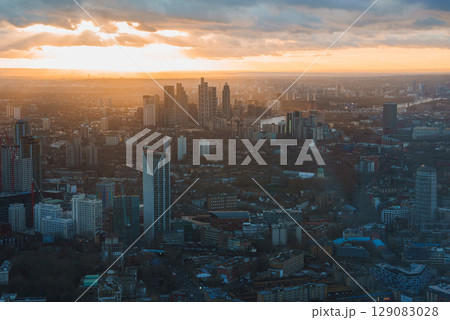 The image shows an aerial view of London's skyline at sunset, featuring modern skyscrapers, iconic buildings, and the River Thames under a warm sky. The image shows an aerial view of London's skyline at sunset, featuring modern skyscrapers, iconic buildings, and the River Thames under a warm sky. 129083028