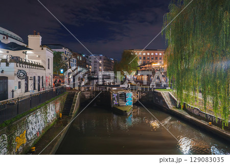 Night scene of Camden Lock in London during Christmas, featuring a canal with festive lights, a bridge, graffiti, and weeping willow trees. 129083035