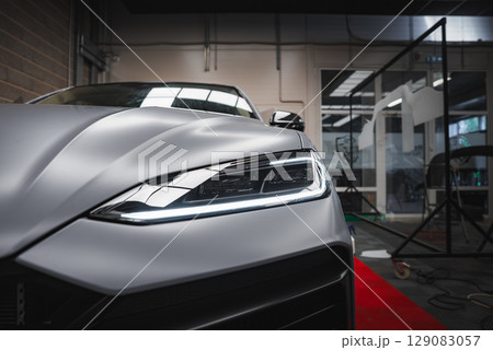Close up view of the Lamborghini Urus front left headlight and hood with matte silver finish, set in an indoor showroom with glass partitions and red carpet. Close up view of the Lamborghini Urus front left headlight and hood with matte silver finish, set in an indoor showroom with glass partitions and red carpet. 129083057