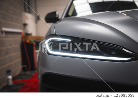 Close up view of the Lamborghini Urus front left headlight with angular LED design, matte silver finish, and red carpet in a garage or showroom. Close up view of the Lamborghini Urus front left headlight with angular LED design, matte silver finish, and red carpet in a garage or showroom. 129083059