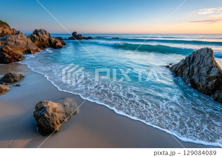 Turquoise ocean waves washing sandy beach at sunrise with rocks 129084089
