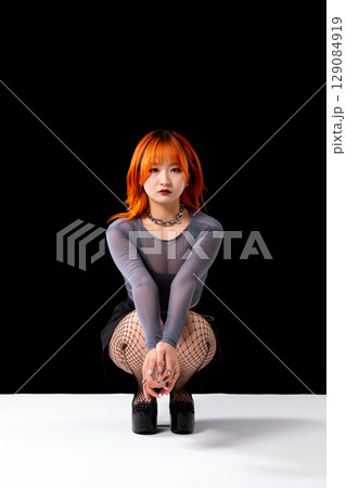 Asian Woman With Orange Hair Squats in Studio Against Black Background Asian Woman With Orange Hair Squats in Studio Against Black Background 129084919
