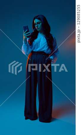 Full-body woman looking at smartphone under blue-orange light Full-body woman looking at smartphone under blue-orange light 129085255