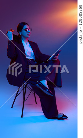 Businesswoman holding smartphone while sitting and playing with her hair under neon light 129085269