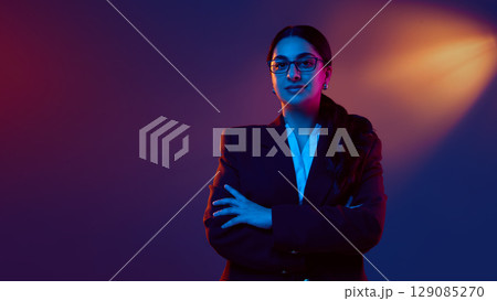 Portrait of confident businesswoman in blazer with crossed arms under neon light 129085270