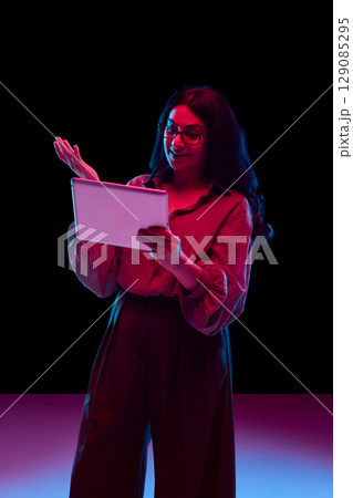 Smiling woman holding tablet and gesturing during video call in neon light 129085295