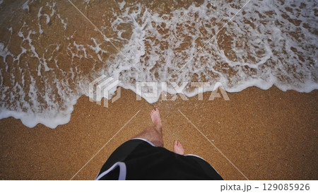 A man in swimming trunks stands barefoot by the sea. The waves are lapping at his feet. 129085926