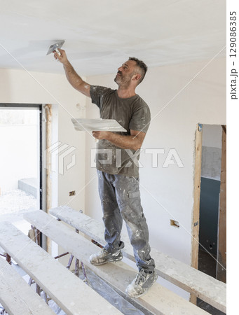 Man working standing on scaffoldings, use trowel and hawk to plaster ceiling, construction 129086385