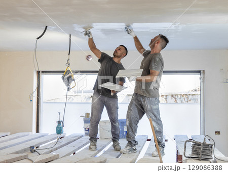 Two men working standing on scaffoldings, use trowel and hawk to plaster ceiling, construction Two men working standing on scaffoldings, use trowel and hawk to plaster ceiling, construction 129086388