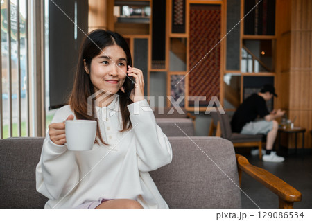 Young woman in a white sweater talking on the phone while holding a coffee cup in a stylish cafe setting. Young woman in a white sweater talking on the phone while holding a coffee cup in a stylish cafe setting. 129086534