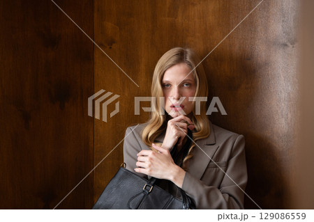 A stylish young woman, long blonde hair, poses in a grey suit, holding a black leather bag against a warm wooden background A stylish young woman, long blonde hair, poses in a grey suit, holding a black leather bag against a warm wooden background 129086559
