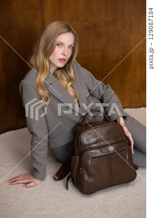 A stylish young woman, long blonde hair, sitting and poses in a grey suit, holding a brown leather backpack against a warm wooden background A stylish young woman, long blonde hair, sitting and poses in a grey suit, holding a brown leather backpack against a warm wooden background 129087184