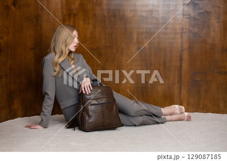 A stylish young woman, long blonde hair, sitting and poses in a grey suit, holding a brown leather backpack against a warm wooden background A stylish young woman, long blonde hair, sitting and poses in a grey suit, holding a brown leather backpack against a warm wooden background 129087185