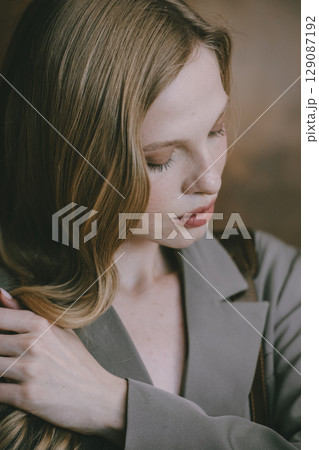 A beautiful young woman with long blonde wavy hair, wearing a stylish grey blazer, gazes thoughtfully to the side against a warm, blurred background A beautiful young woman with long blonde wavy hair, wearing a stylish grey blazer, gazes thoughtfully to the side against a warm, blurred background 129087192