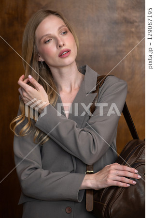 A stylish young woman, long blonde hair, poses in a grey suit, holding a brown leather backpack against a warm wooden background 129087195